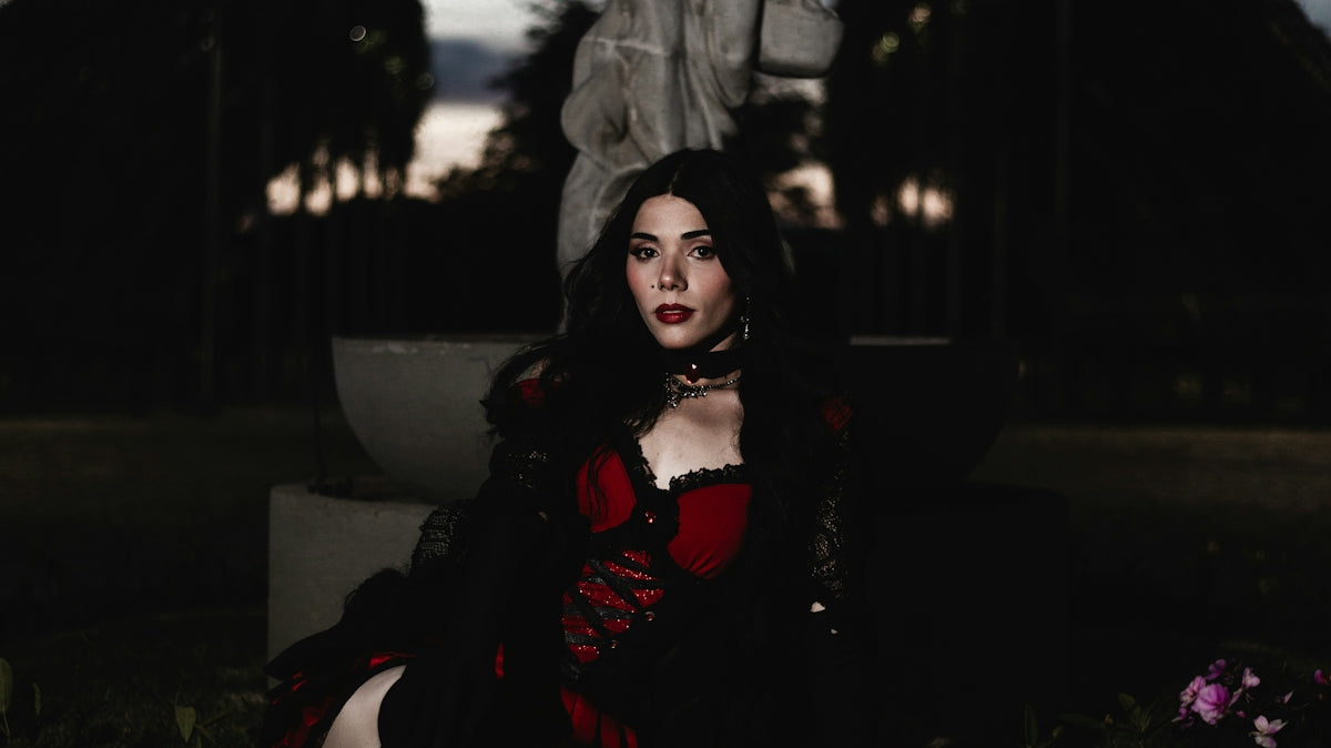 Woman in gothic attire sits by statue at dusk