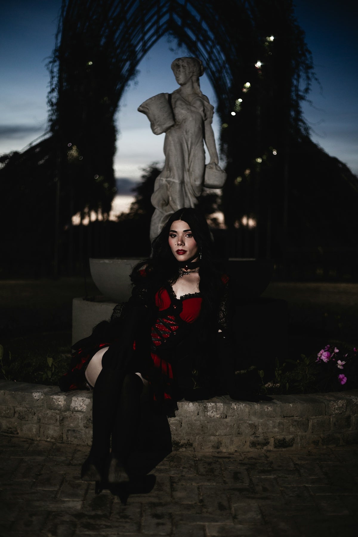 Woman in gothic attire sits by statue at dusk